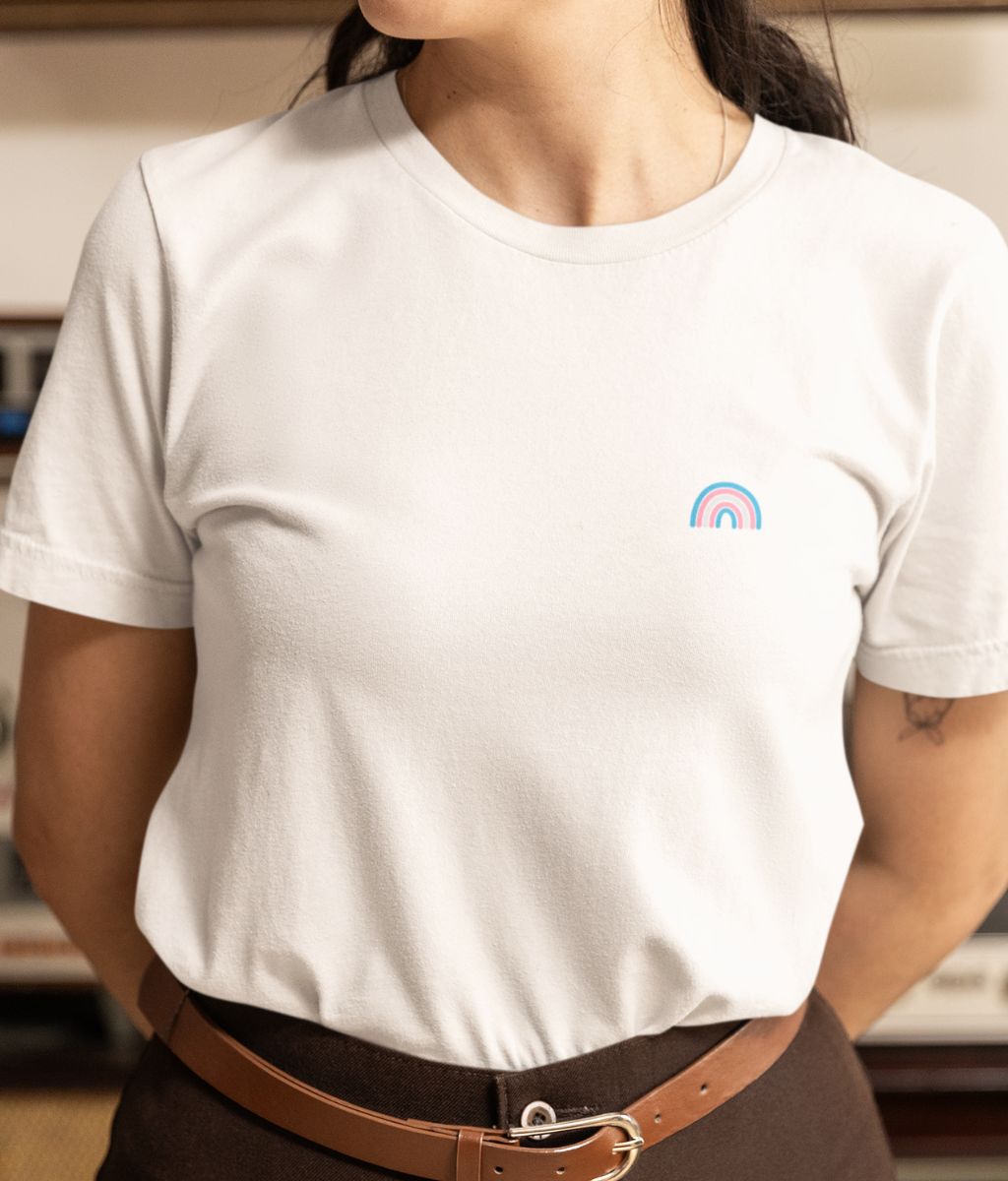 A woman in a white t-shirt featuring a trans rainbow logo, smiling and standing against a neutral background.
